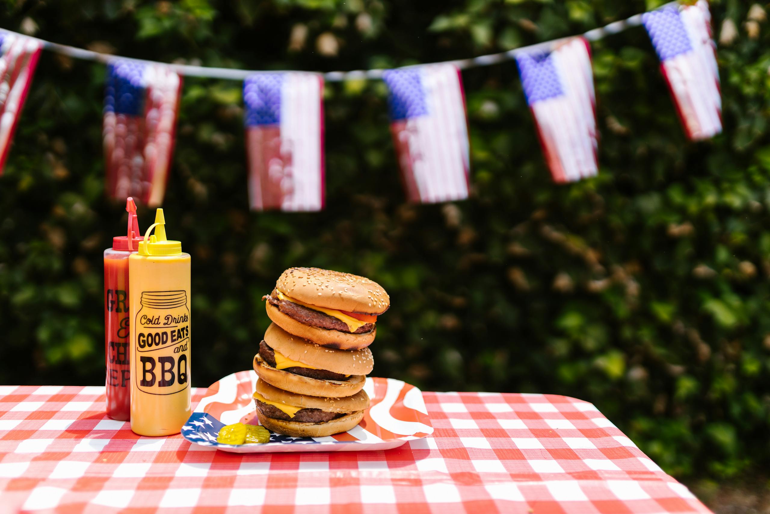 Stacked burgers on a table with American flag decor and condiments. Perfect for festive outdoor BBQs.